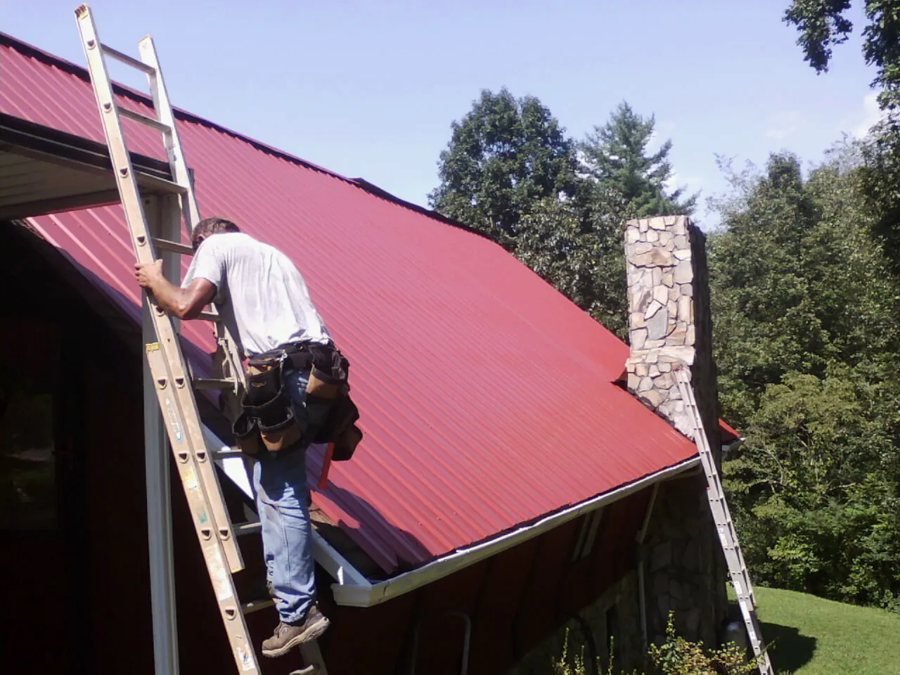Roofer on a ladder beside a red metal roof and stone chimney — Soffit & Fascia Repair by Roofing Brothers in Baldwin City